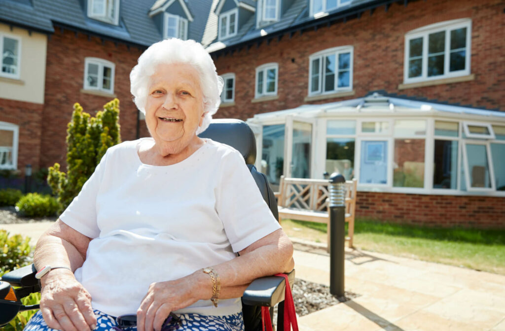 A smiling older adult woman sitting on a wheelchair looking directly at the camera against a memory care facility