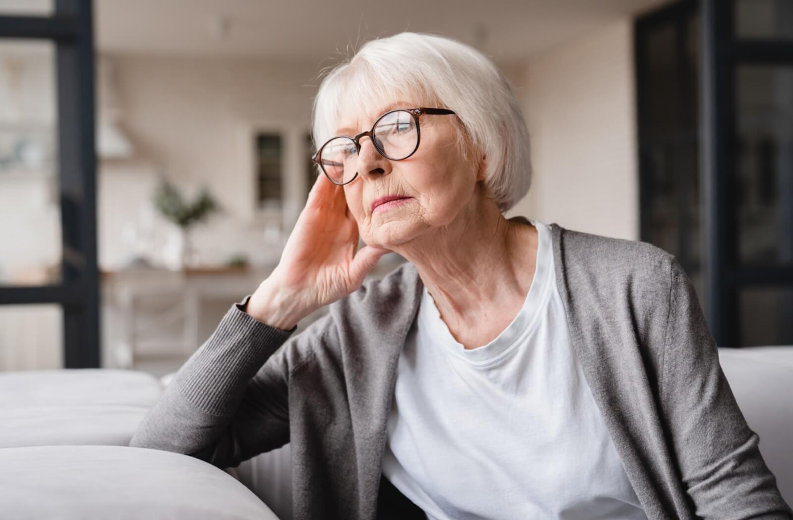 An older adult woman with glasses on sitting on a couch and looking out the window with a serious expression