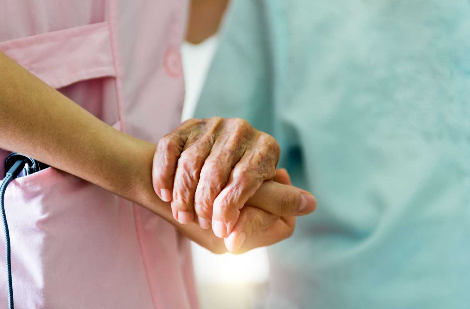 A close-up image of a nurse in pink scrubs holding the hand of a senior resident.