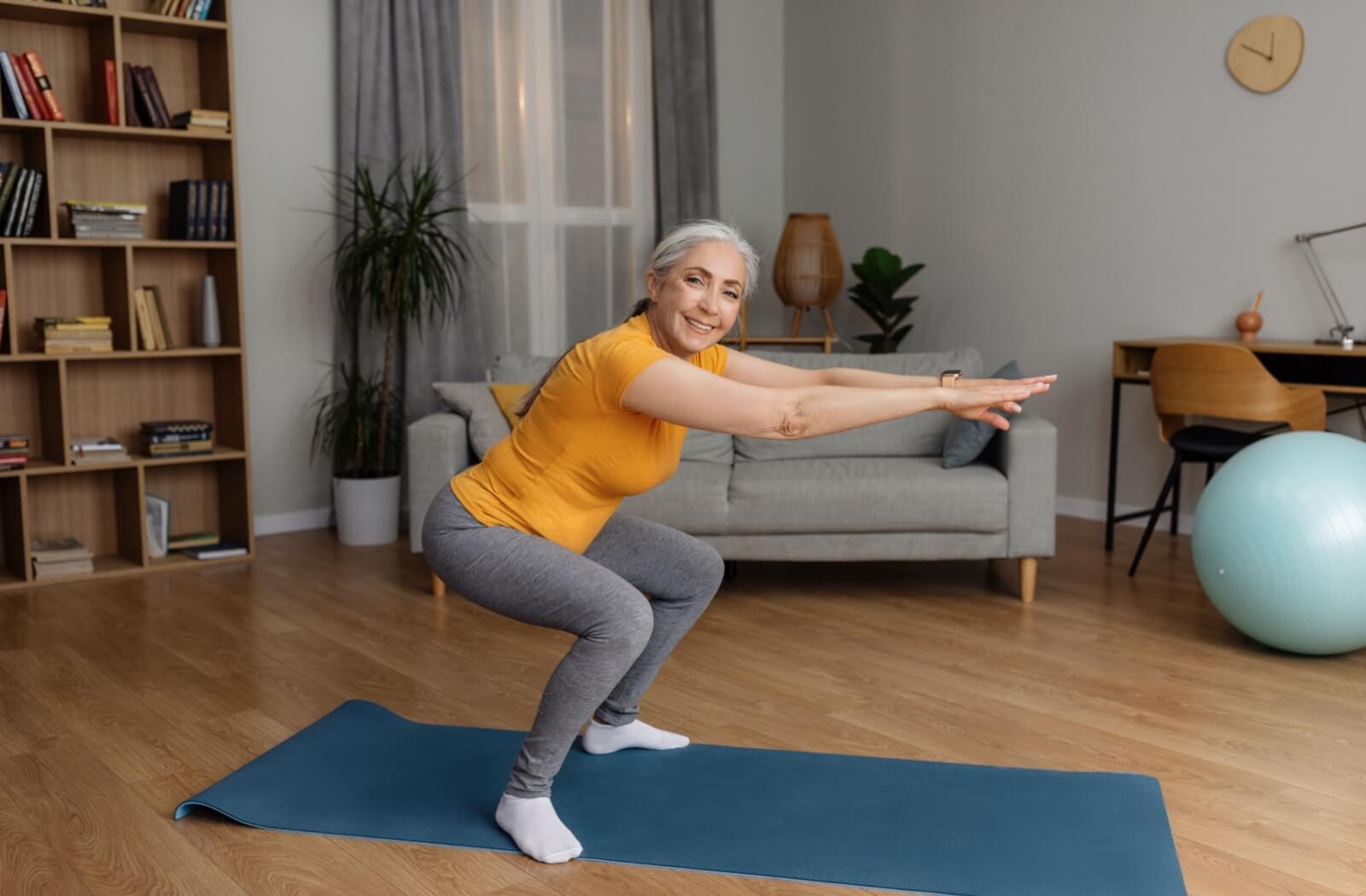 A smiling senior performs a bodyweight squat from the comfort of their senior living community home.